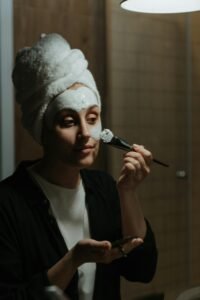 A woman in a towel applies a face mask as part of her skincare routine in a cozy setting.