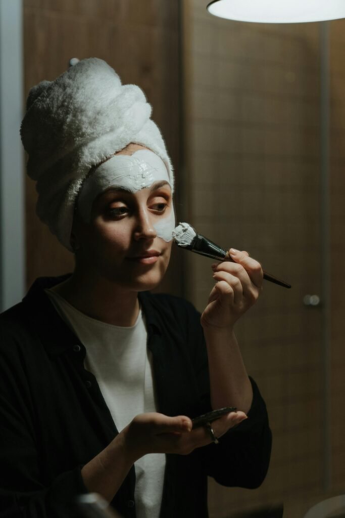 A woman in a towel applies a face mask as part of her skincare routine in a cozy setting.